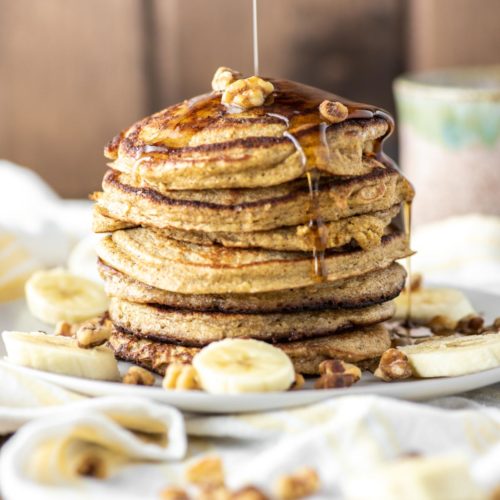 banana oat pancakes stacked on plate with maple syrup being drizzled on top