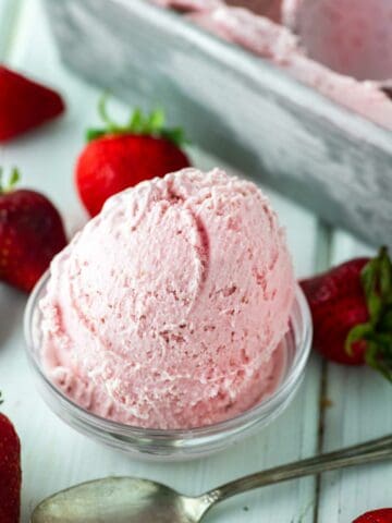 strawberry ice cream in glass bowl with strawberries around it and metal tin behind it