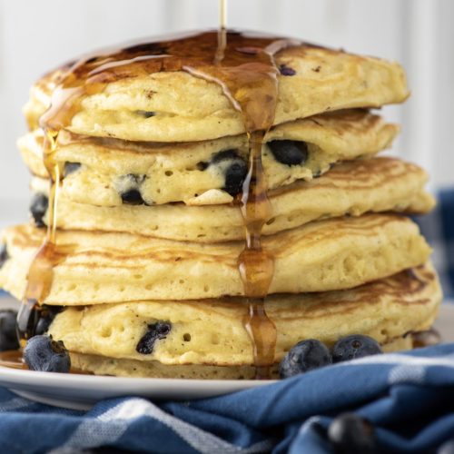 maple syrup being drizzled over stacked blueberry pancakes
