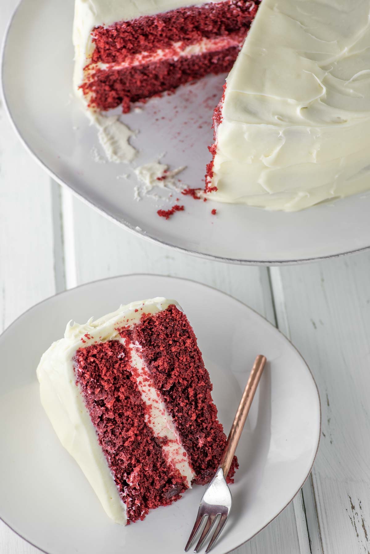 slice of red velvet cake on white plate with fork with cake stand in background