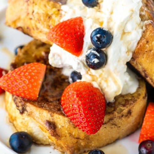 3 slices of sourdough French toast on white plate with whipped cream, strawberries and blueberries