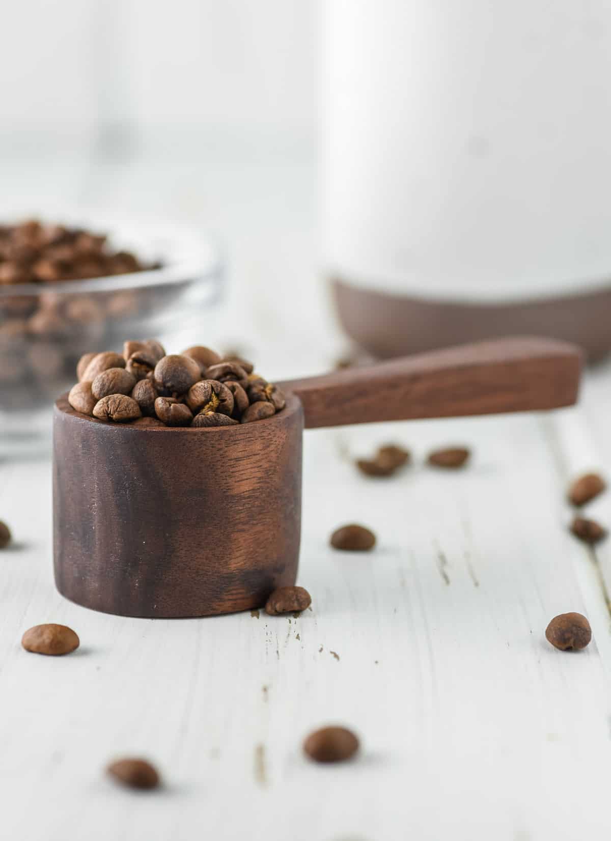 handmade coffee scoop with coffee beans on white board with bowl of coffee beans and mug in background