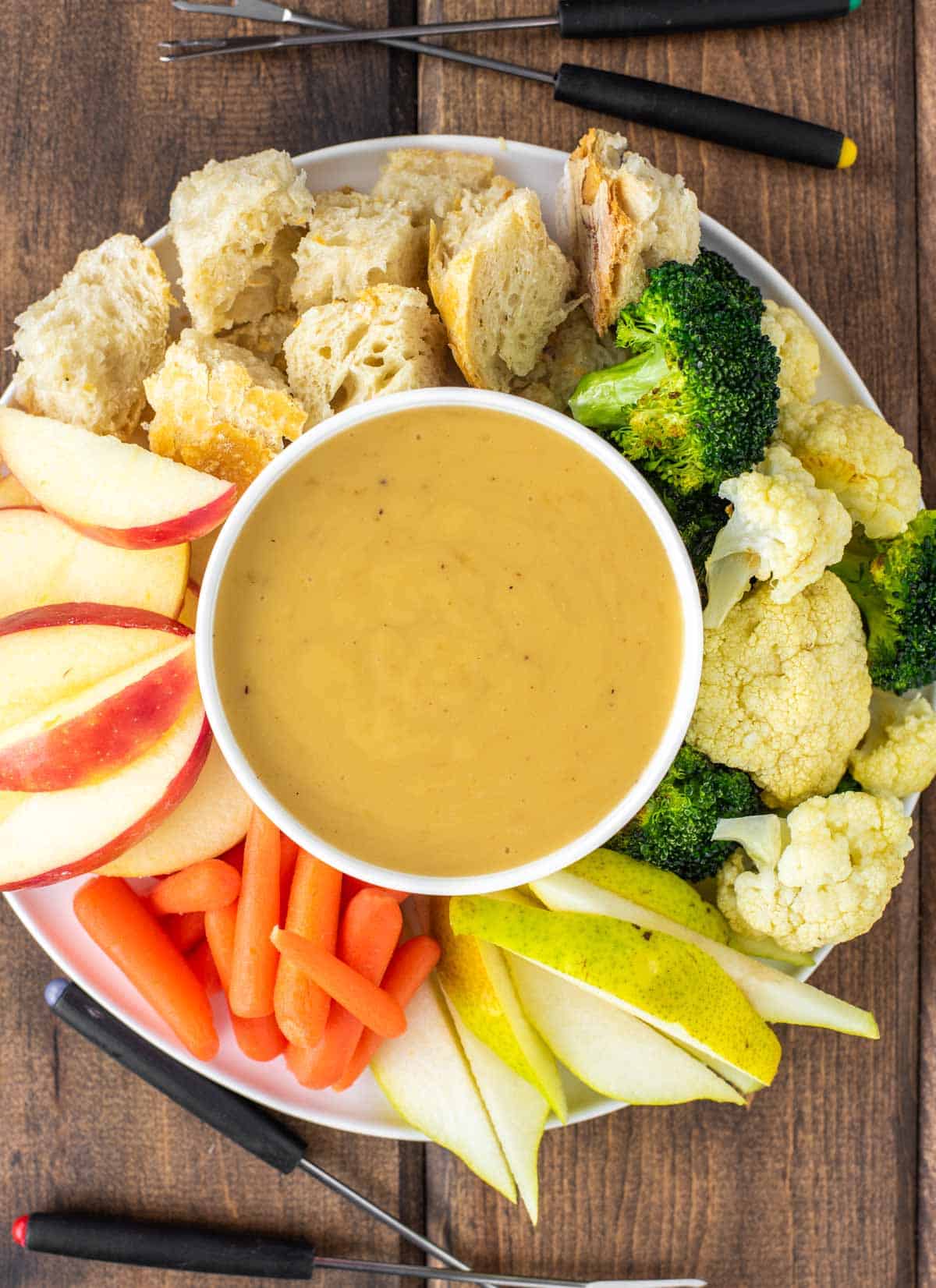 overhead shot of fondue in white bowl with plate of veggies, fruit and bread
