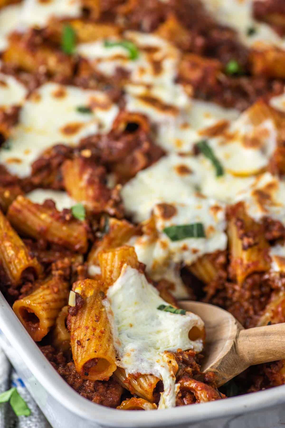 close-up of wooden spoon dipped in white baking dish of rigatoni al forno