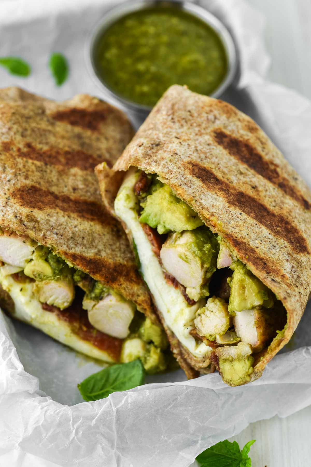 chicken pesto wrap stacked on parchment-lined sheet with bowl of pesto behind it