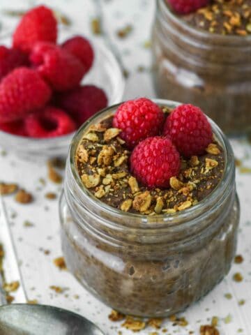 two mason jars of chocolate chia seed pudding with glass of raspberries in background