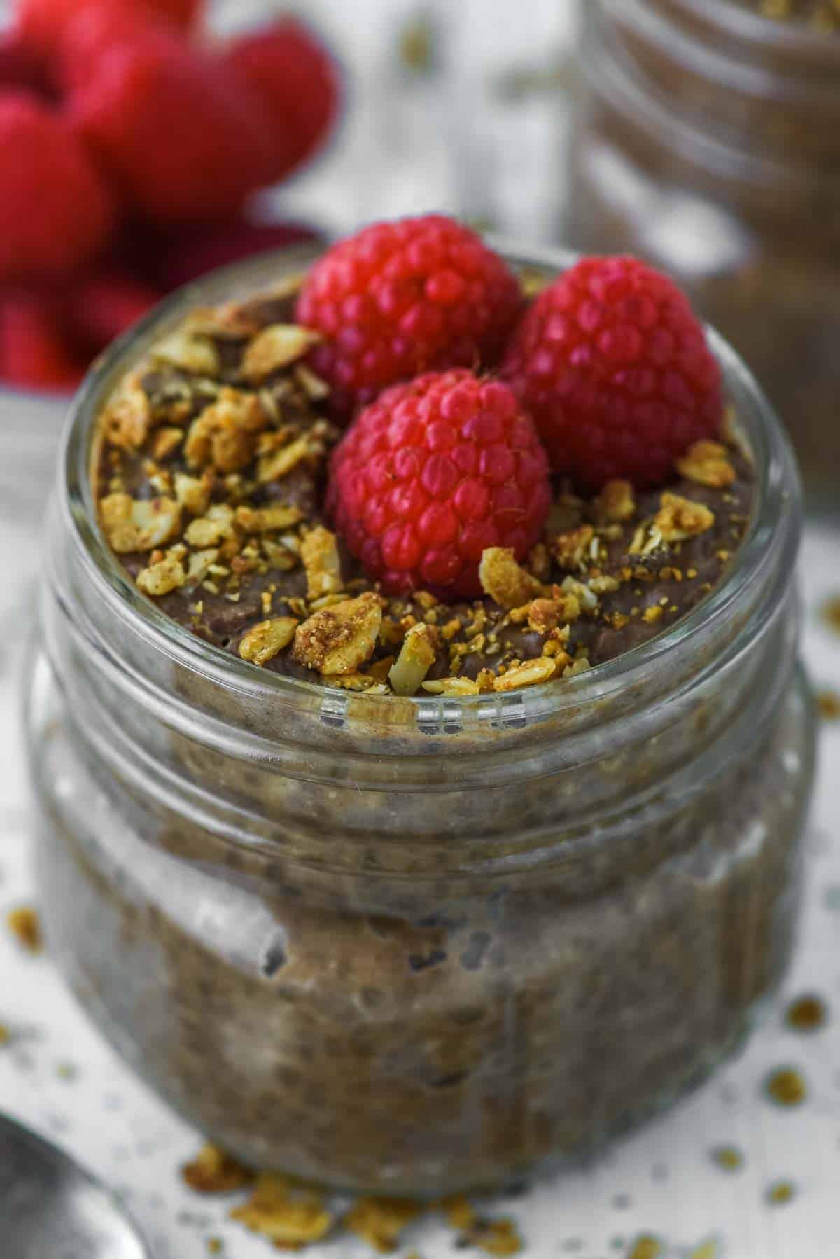 close-up of chocolate chia seed pudding in glass jar with raspberries and granola on top